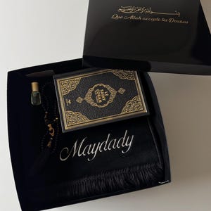 K&ouml;nnte beinhalten: Eine schwarze Geschenkbox mit einem schwarz-goldenen Le Saint Coran Buch, einem schwarzen Gebetsteppich mit dem in Wei&szlig; gestickten Namen Maydady, einer schwarzen Gebetskette und einem kleinen Glasparf&uuml;mfl&auml;schchen. Der Deckel tr&auml;gt goldenen Text.
