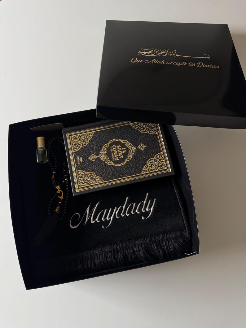 K&ouml;nnte beinhalten: Eine schwarze Geschenkbox mit einem schwarz-goldenen Le Saint Coran Buch, einem schwarzen Gebetsteppich mit dem in Wei&szlig; gestickten Namen Maydady, einer schwarzen Gebetskette und einem kleinen Glasparf&uuml;mfl&auml;schchen. Der Deckel tr&auml;gt goldenen Text.