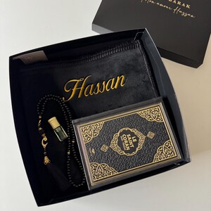 K&ouml;nnte beinhalten: Eine schwarze Geschenkbox mit einem schwarzen Gebetsteppich, der den Namen Hassan in Gold gestickt hat, einer schwarzen Gebetskette mit goldenem Halbmondanh&auml;nger, einem kleinen Glasparf&uuml;mfl&auml;schchen und einem schwarzen Buch namens Le Saint Coran.