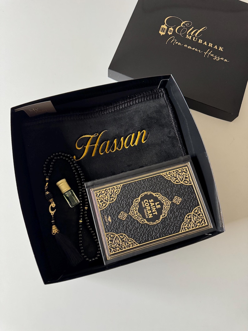 K&ouml;nnte beinhalten: Eine schwarze Geschenkbox mit einem schwarzen Gebetsteppich, der den Namen Hassan in Gold gestickt hat, einer schwarzen Gebetskette mit goldenem Halbmondanh&auml;nger, einem kleinen Glasparf&uuml;mfl&auml;schchen und einem schwarzen Buch namens Le Saint Coran.