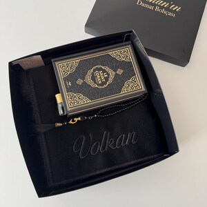 K&ouml;nnte beinhalten: Eine schwarze Geschenkbox mit einem schwarzen Gebetsteppich, der mit dem Namen Volkan bestickt ist, einem Buch mit dem Titel Le Saint Coran mit goldenen Zierr&auml;ndern, einem kleinen Glasparf&uuml;mfl&auml;schchen mit Goldkappe und einer schwarzen Gebetskette mit Goldquaste.