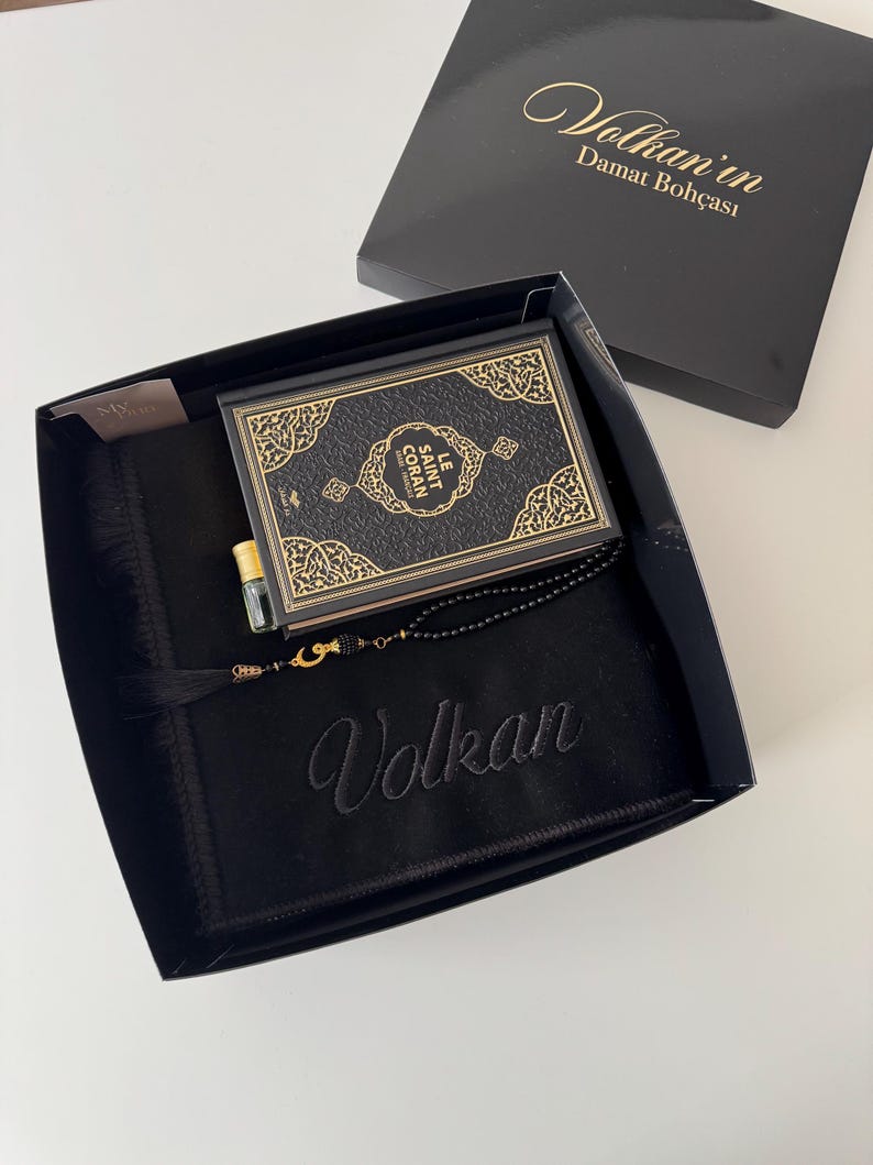 K&ouml;nnte beinhalten: Eine schwarze Geschenkbox mit einem schwarzen Gebetsteppich, der mit dem Namen Volkan bestickt ist, einem Buch mit dem Titel Le Saint Coran mit goldenen Zierr&auml;ndern, einem kleinen Glasparf&uuml;mfl&auml;schchen mit Goldkappe und einer schwarzen Gebetskette mit Goldquaste.