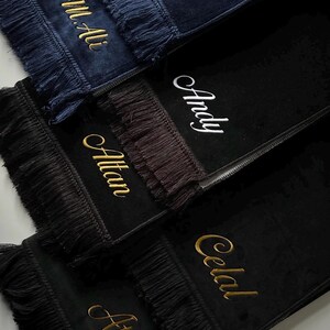 May include: A collection of rectangular prayer mats in navy blue and black velvet, each featuring decorative fringe on the ends. The mats are personalized with embroidered names in gold or white script, including M.Ali, Andy, Altan, Celal, and Atilla.