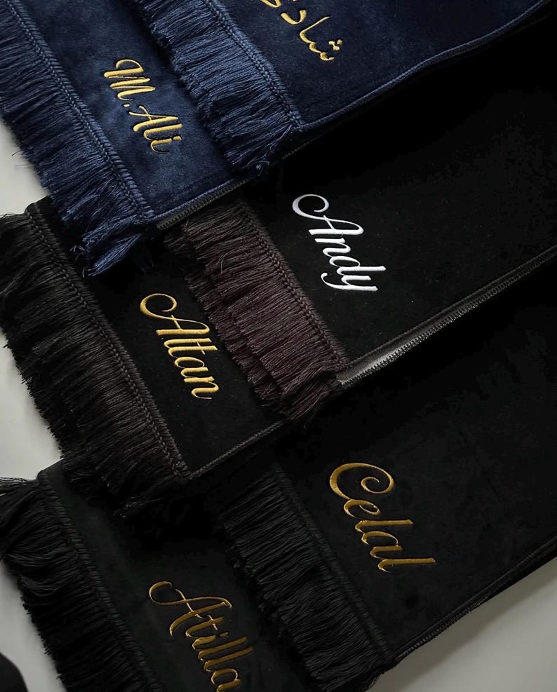 May include: A collection of rectangular prayer mats in navy blue and black velvet, each featuring decorative fringe on the ends. The mats are personalized with embroidered names in gold or white script, including M.Ali, Andy, Altan, Celal, and Atilla.