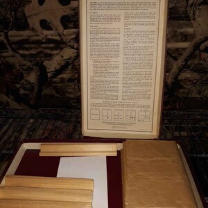 May include: A vintage Scrabble game box with a red lid and a brown interior. The box contains a set of wooden tiles, a rule book, and a paper score sheet. The rule book has the title "Rules for Playing Scrabble" printed on the front.