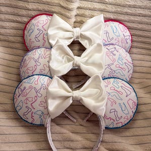 May include: Three mouse ear headbands. Each has a white satin bow and circular ears patterned with a race track design in pink, blue, and purple. The ear edges are trimmed in red, blue, and pink. The headband is white with a matching pattern.