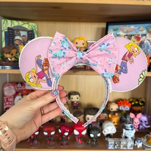 Peut inclure: Serre-tête oreilles de souris rose avec un nœud floral et des illustrations de personnages de dessins animés. Les oreilles sont rose clair avec une bordure en dentelle blanche. Le nœud est rose avec des motifs floraux bleus. Un accessoire amusant.
