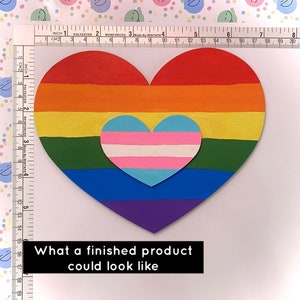 May include: A large heart-shaped cutout painted with horizontal rainbow stripes, featuring a smaller heart-shaped cutout in the centre with horizontal light blue, pink, and white stripes. A ruler shows the large heart is 20 cm wide and 18 cm tall.