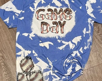 Könnte beinhalten: Ein blaues T-Shirt mit einem weißen Bleichmittel-Design. Das Design zeigt die Worte "GAME DAY" in weißen Buchstaben mit einem Leopardenmuster und Baseballnähten. In der linken unteren Ecke befinden sich drei Baseballs mit Leopardenmuster und Nähten.