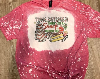 Könnte beinhalten: Rotes T-Shirt mit weißen Bleichmittelspritzern und einer weihnachtlichen Grafik mit dem Text "Torn between lookin' like a snack and eatin' one".