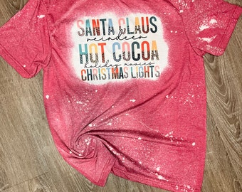 Könnte beinhalten: Rotes T-Shirt mit einem weißen, mit Bleichmittel bespritzten Design. Das Shirt hat eine festliche Weihnachtsgrafik mit dem Text "Santa Claus reindeer hot cocoa holiday movies Christmas lights".