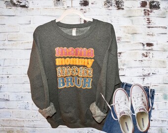 Könnte beinhalten: Ein dunkelgrauer Pullover mit dem Text "mama mommy mom bruh" in bunten Retro-Lettern. Der Pullover liegt auf einem weißen Ziegelhintergrund mit einem Paar weißen Canvas-Schuhen und einer blauen Jeans.