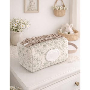 May include: A rectangular quilted fabric pouch featuring a light green floral pattern on an off-white background. The pouch includes a beige ruffled zipper closure, a side handle, and a white plastic flip-top dispenser lid centered on the front.