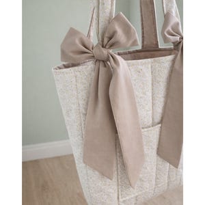May include: A quilted tote bag featuring a light beige floral pattern on a cream background. The bag includes two long shoulder straps, each adorned with a large, solid taupe-colored fabric bow tied at the base of the strap attachment point.