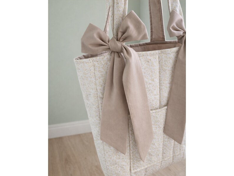 May include: A quilted tote bag featuring a light beige floral pattern on a cream background. The bag includes two long shoulder straps, each adorned with a large, solid taupe-colored fabric bow tied at the base of the strap attachment point.