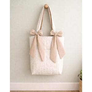 May include: A quilted tote bag in a light cream color with a subtle pattern. The bag features two long shoulder straps, each adorned with a large, tied beige ribbon bow at the base where the strap meets the top edge of the rectangular bag body.