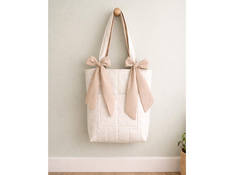May include: A quilted tote bag in a light cream color with a subtle pattern. The bag features two long shoulder straps, each adorned with a large, tied beige ribbon bow at the base where the strap meets the top edge of the rectangular bag body.