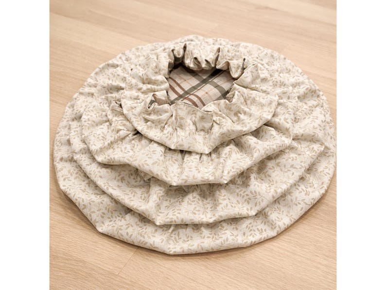 May include: A set of three nested circular fabric bowl covers featuring a light beige background with a small repeating leaf pattern. The covers have elasticized edges for a snug fit, and the innermost cover reveals a brown and white plaid lining.