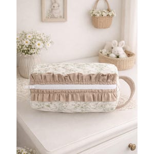 May include: A rectangular quilted cosmetic bag featuring a light cream fabric with a subtle green floral pattern. The bag includes a white zipper closure flanked by two rows of beige ruffles and a matching beige side handle for easy carrying.