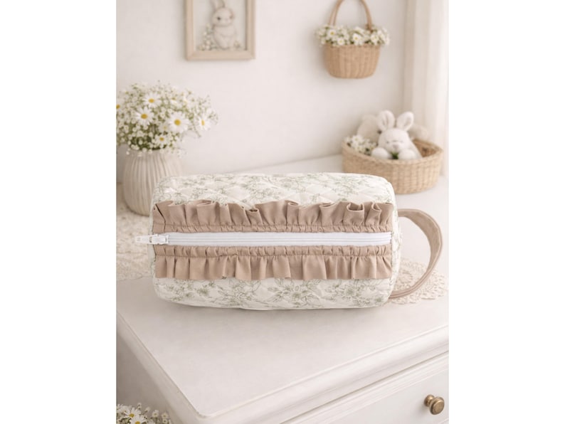 May include: A rectangular quilted cosmetic bag featuring a light cream fabric with a subtle green floral pattern. The bag includes a white zipper closure flanked by two rows of beige ruffles and a matching beige side handle for easy carrying.