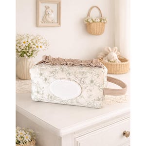 May include: A rectangular fabric wipes case featuring a light beige floral pattern on a white background. The top edge is decorated with a ruffled beige trim, and the front includes a white plastic flip-top lid. A beige handle is attached to the side.