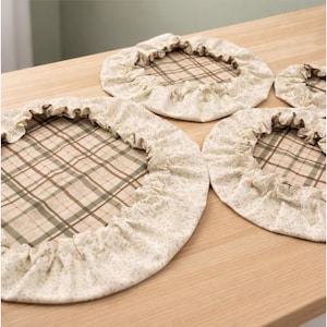 May include: A set of three round fabric bowl covers with elasticized ruffled edges. The center of each cover features a tan and green plaid pattern, while the ruffled border is made of a cream-colored fabric with a subtle floral print.