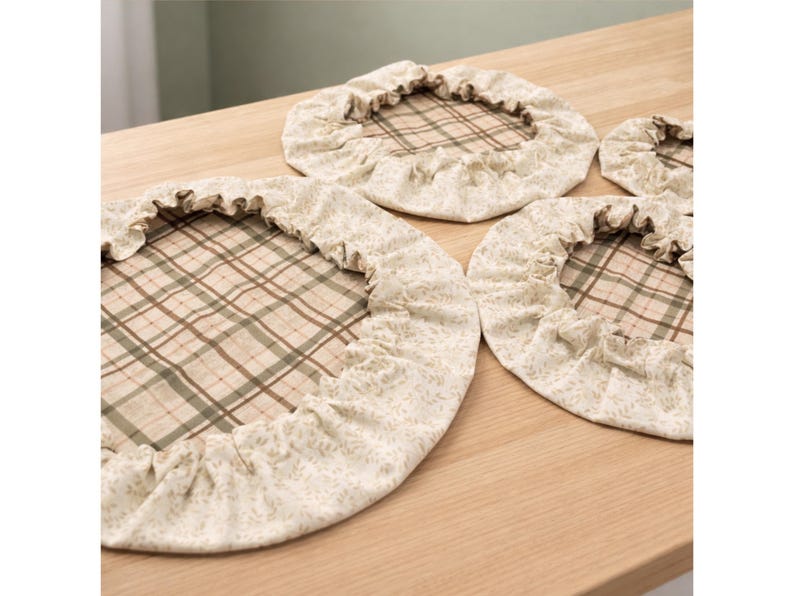 May include: A set of three round fabric bowl covers with elasticized ruffled edges. The center of each cover features a tan and green plaid pattern, while the ruffled border is made of a cream-colored fabric with a subtle floral print.