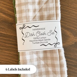 May include: A set of dish cloths featuring a beige and white gingham pattern on one side and a green fabric with white paw prints on the other. A white paper band wraps around the cloths with the text HANDMADE Dish Cloth Set 100% Cotton.