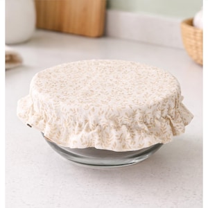 May include: A round glass bowl covered with a beige fabric lid featuring a small repeating leaf pattern. The fabric cover has an elasticized, ruffled edge that fits snugly over the rim of the bowl, securing the top in a gathered, decorative style.