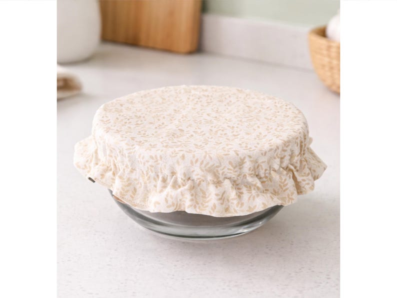 May include: A round glass bowl covered with a beige fabric lid featuring a small repeating leaf pattern. The fabric cover has an elasticized, ruffled edge that fits snugly over the rim of the bowl, securing the top in a gathered, decorative style.