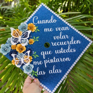 May include: A blue graduation cap decorated with white butterflies, yellow and blue paper flowers, and rhinestones. The cap has the text "Cuando me vean volar recuerden que ustedes me pintaron las alas" written in white.