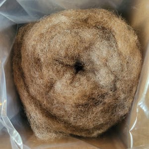 May include: A circular coil of light brown, fibrous material with a coarse, tangled texture. The fibers are densely packed into a round shape with a small, dark, hollow center, contained within a clear plastic bag inside a cardboard box.
