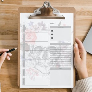 May include: A clipboard with a weekly planner featuring a skull and floral design. The planner includes sections for priorities, to-do lists, and notes. A pencil is held near the planner.