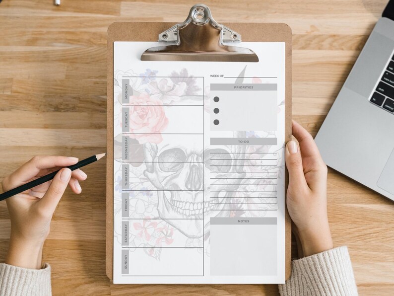 May include: A clipboard with a weekly planner featuring a skull and floral design. The planner includes sections for priorities, to-do lists, and notes. A pencil is held near the planner.