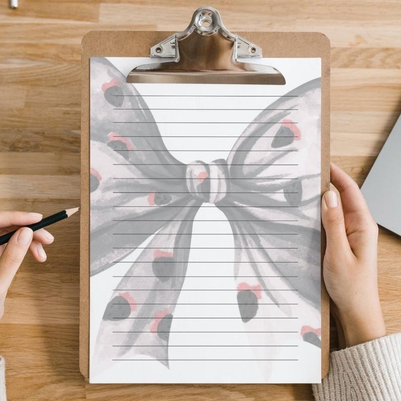 Peut inclure: Un presse-papiers avec une feuille de papier lign&eacute; orn&eacute;e d'un motif de n&oelig;ud papillon &agrave; l'aquarelle gris et rouge. Le presse-papiers a un clip en m&eacute;tal argent&eacute; et un dos marron. Une main tient un crayon, pr&ecirc;t &agrave; &eacute;crire.