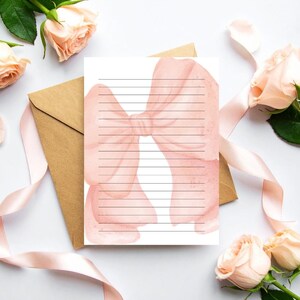 Peut inclure: Une feuille de papeterie lign&eacute;e avec un motif de n&oelig;ud rose &agrave; l'aquarelle. Une enveloppe marron, un ruban rose et des roses p&ecirc;che sont dispos&eacute;s autour de la papeterie sur une surface blanche. Parfait pour &eacute;crire des lettres.
