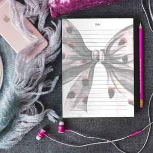 Peut inclure: Un bloc-notes lign&eacute; avec un motif de n&oelig;ud papillon &agrave; l'aquarelle gris et rose. Le mot &laquo; Notes &raquo; est en haut. Un crayon rose, un t&eacute;l&eacute;phone, une &eacute;charpe et des &eacute;couteurs sont dispos&eacute;s autour du bloc-notes.