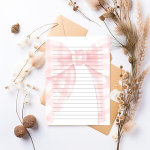 Peut inclure: Une feuille de papier &agrave; lettres lign&eacute;e avec un motif de n&oelig;ud rose. Le n&oelig;ud a un motif vichy et se trouve en haut au centre de la page. La papeterie est sur une surface blanche avec des fleurs s&eacute;ch&eacute;es et une enveloppe marron.