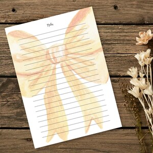 Peut inclure: Bloc-notes lign&eacute; avec un motif de n&oelig;ud aquarelle dans les tons p&ecirc;che et jaune. Le mot "Notes" est &eacute;crit en haut. Le bloc-notes est pos&eacute; sur une surface en bois avec des fleurs s&eacute;ch&eacute;es.