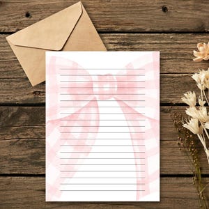 Peut inclure: Papier &agrave; lettres lign&eacute; avec un motif de n&oelig;ud rose &agrave; carreaux. Une enveloppe marron est en arri&egrave;re-plan, ainsi que des fleurs s&eacute;ch&eacute;es, le tout sur une surface en bois. Parfait pour &eacute;crire des lettres.