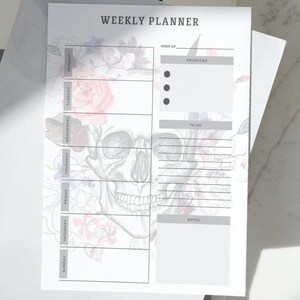 May include: A white weekly planner with a skull and floral design. The planner includes sections for each day of the week, priorities, to-do lists, and notes. The text "WEEKLY PLANNER" is at the top.