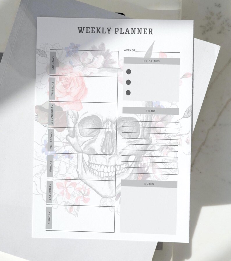 May include: A white weekly planner with a skull and floral design. The planner includes sections for each day of the week, priorities, to-do lists, and notes. The text "WEEKLY PLANNER" is at the top.