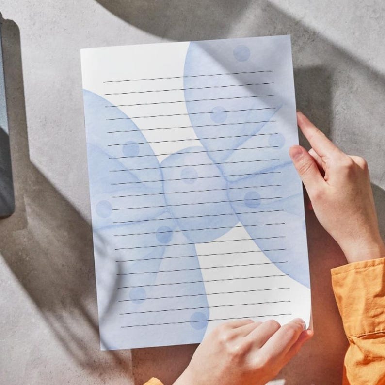 Peut inclure: Une feuille de papeterie lign&eacute;e avec un motif aquarelle de cercles bleus doux. Le papier est tenu par des mains, avec une manche orange visible. Le papier comporte des lignes horizontales pour l'&eacute;criture.