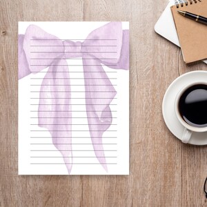 Peut inclure: Une feuille de papier &agrave; lettres lign&eacute;e avec une illustration &agrave; l'aquarelle d'un n&oelig;ud lavande en haut. Les rubans du n&oelig;ud descendent sur la page. Le papier est sur un bureau en bois, avec une tasse de caf&eacute;, un carnet et un stylo en arri&egrave;re-plan.