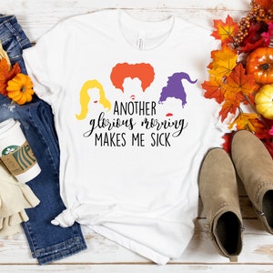 May include: White t-shirt with the phrase "ANOTHER glorious morning MAKES ME SICK" and silhouettes of three witches. The shirt is styled with blue jeans, fall foliage, a coffee cup, and tan boots. A seasonal, fall-themed design.