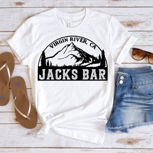 May include: White t-shirt with a black graphic featuring a mountain range, trees, and the text "Virgin River, CA" above "Jacks Bar". The shirt is paired with brown sandals, denim shorts, and sunglasses.