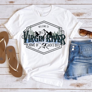 May include: White t-shirt with a graphic that reads "Welcome to Virgin River, Home of Jack's Bar." The design features mountains, trees, and a river scene. The shirt is paired with denim shorts, sunglasses, and brown sandals.