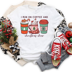 May include: White t-shirt with the text "I run on coffee and Christmas cheer" and a graphic of three festive coffee cups. The cups are red, green, and pink, decorated with Christmas trees, snowflakes, and candy canes. Red sneakers and Christmas ornaments are also in the image.