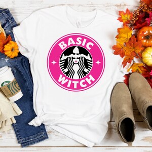 以下が含まれることがあります： 「BASIC WITCH」と書かれたピンクと黒のグラフィックが特徴の白いTシャツ。青いジーンズ、コーヒーカップ、スカーフ、カボチャや葉など秋をテーマにした装飾が施されています。ベージュのアンクルブーツがアンサンブルを完成させています。