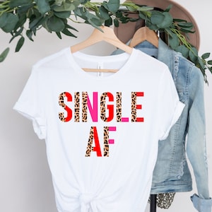 May include: White t-shirt with the text "SINGLE AF" in a bold, colorful design. The letters are a mix of red, pink, and leopard print. The shirt is hanging on a wooden hanger, with a denim jacket and hat in the background.